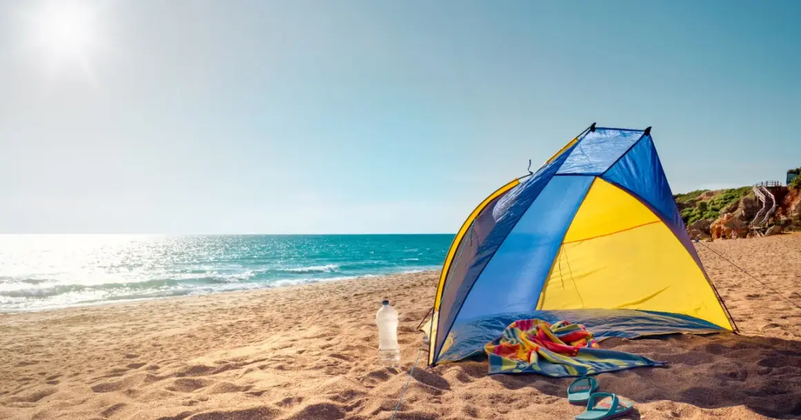 Kolorowy namiot na plaży, obok butelka wody, ręcznik i klapki. Słońce odbija się w morzu.