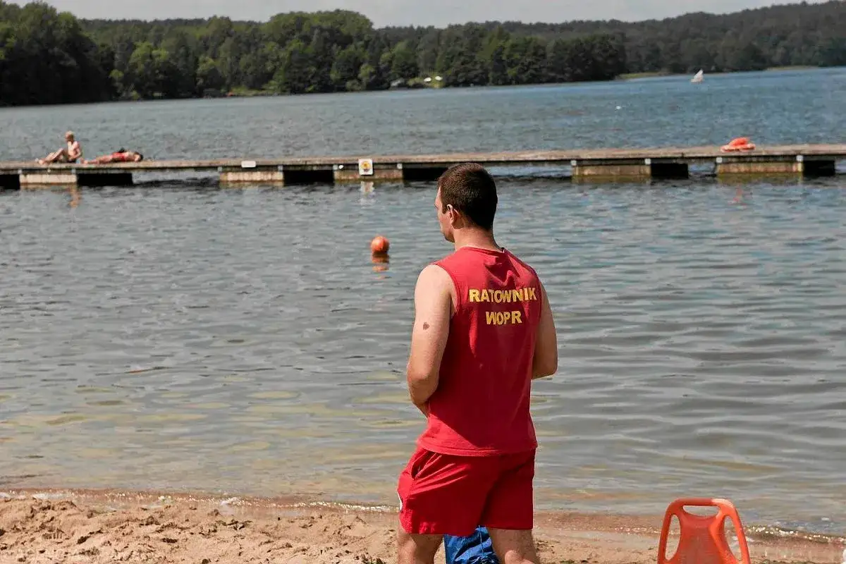 Ratownik WOPR czuwa nad bezpieczeństwem podczas pływania w jeziorze. Na pomoście leżą plażowicze.